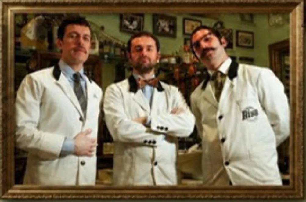Tre barbieri di BisB in giacca bianca col collo nero, davanti alle specchiere della bottega; a sinistra Andrea Cottone